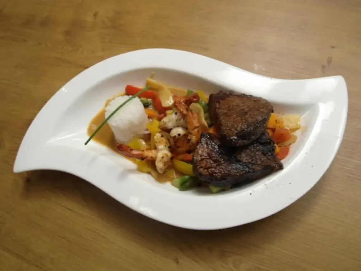 Zwei saftige Rinderfiletstücke auf einem Bett aus Gemüse und Garnelen, serviert mit Reis in einer cremigen Sauce, die an einen **thai curry** erinnert.