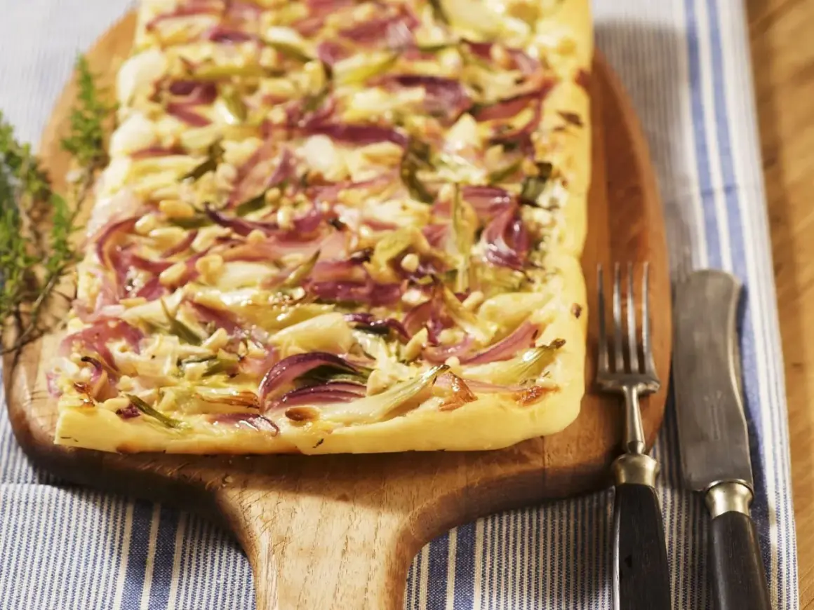 Flammkuchen mit roten Zwiebeln, Pinienkernen und Käse auf einem Holzbrett, daneben Gabel und Messer.