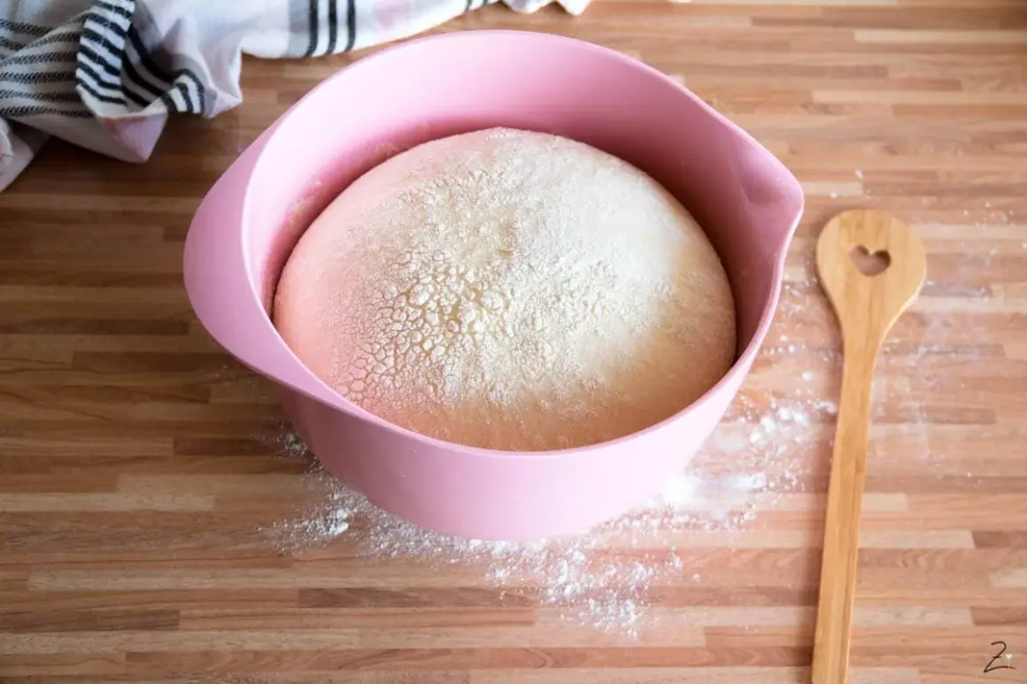Hefeteig für einen Blechkuchen ruht in einer rosa Schüssel. Daneben liegt ein Holzlöffel mit Herz.
