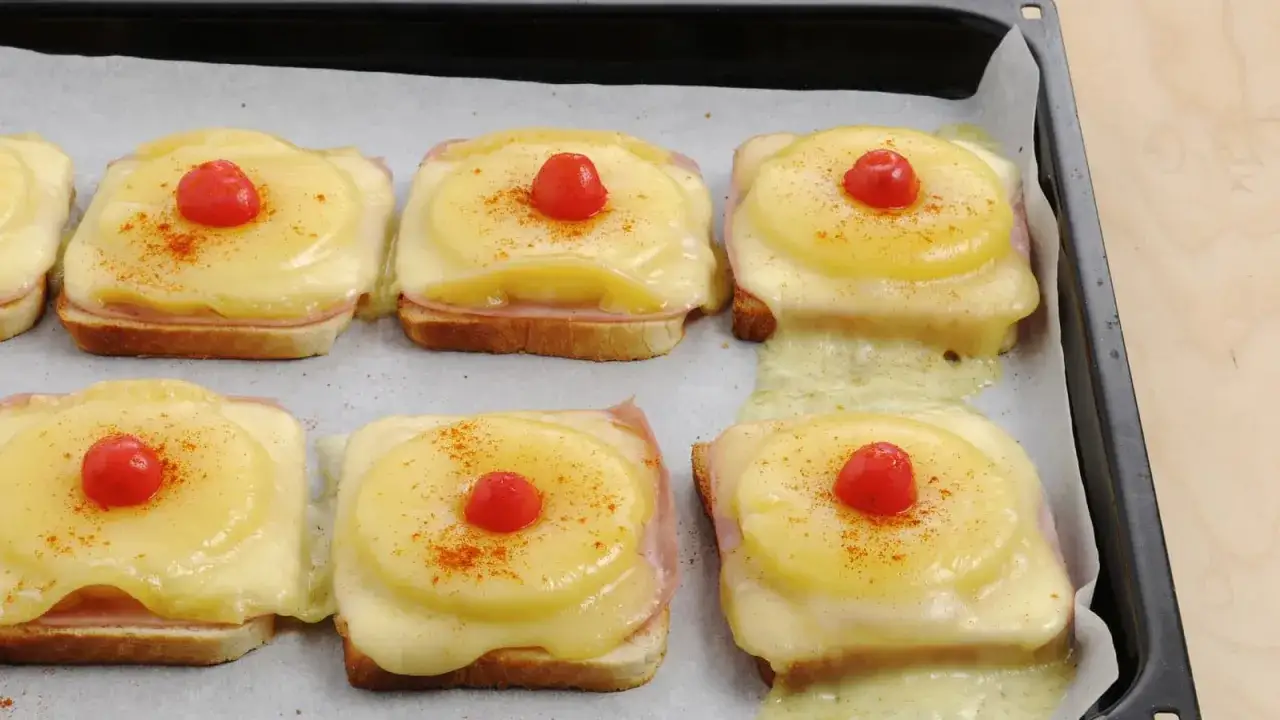 Sechs &uuml;berbackene Toast Hawaii mit Schinken, Ananas, K&auml;se und Kirsche auf einem Backblech.