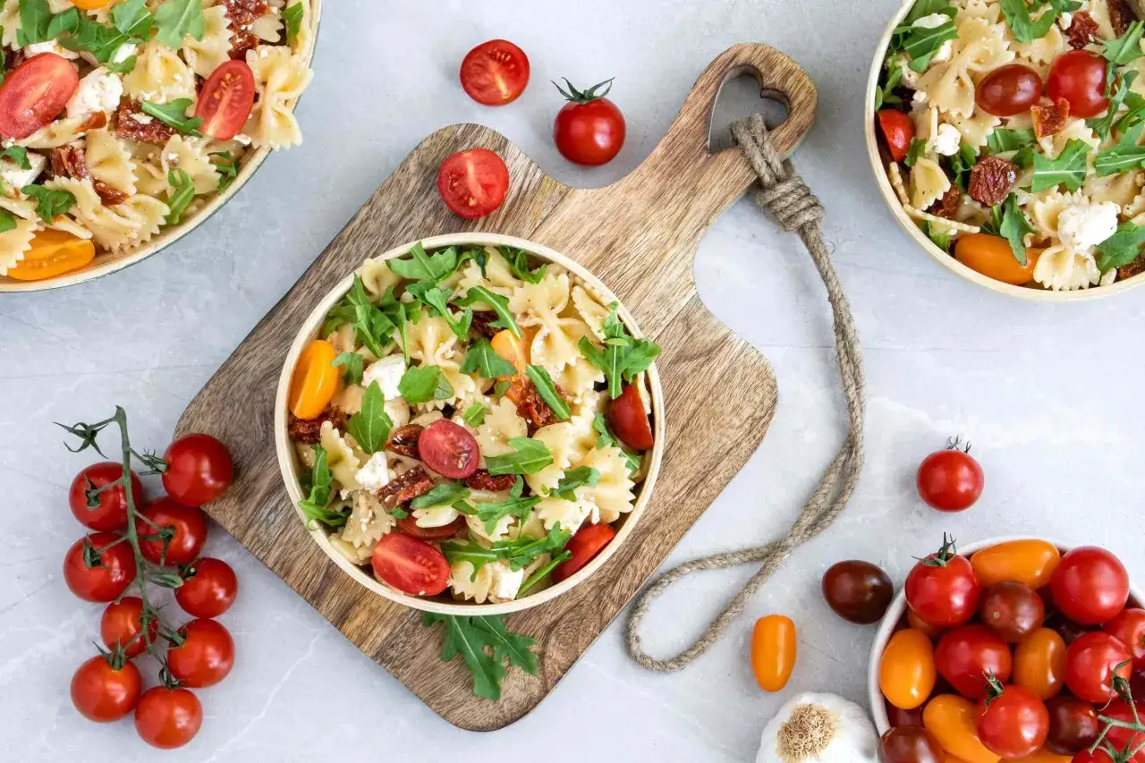 Ein köstlicher mediterraner Nudelsalat mit Farfalle, Kirschtomaten, Rucola und Feta auf einem Holzbrett.