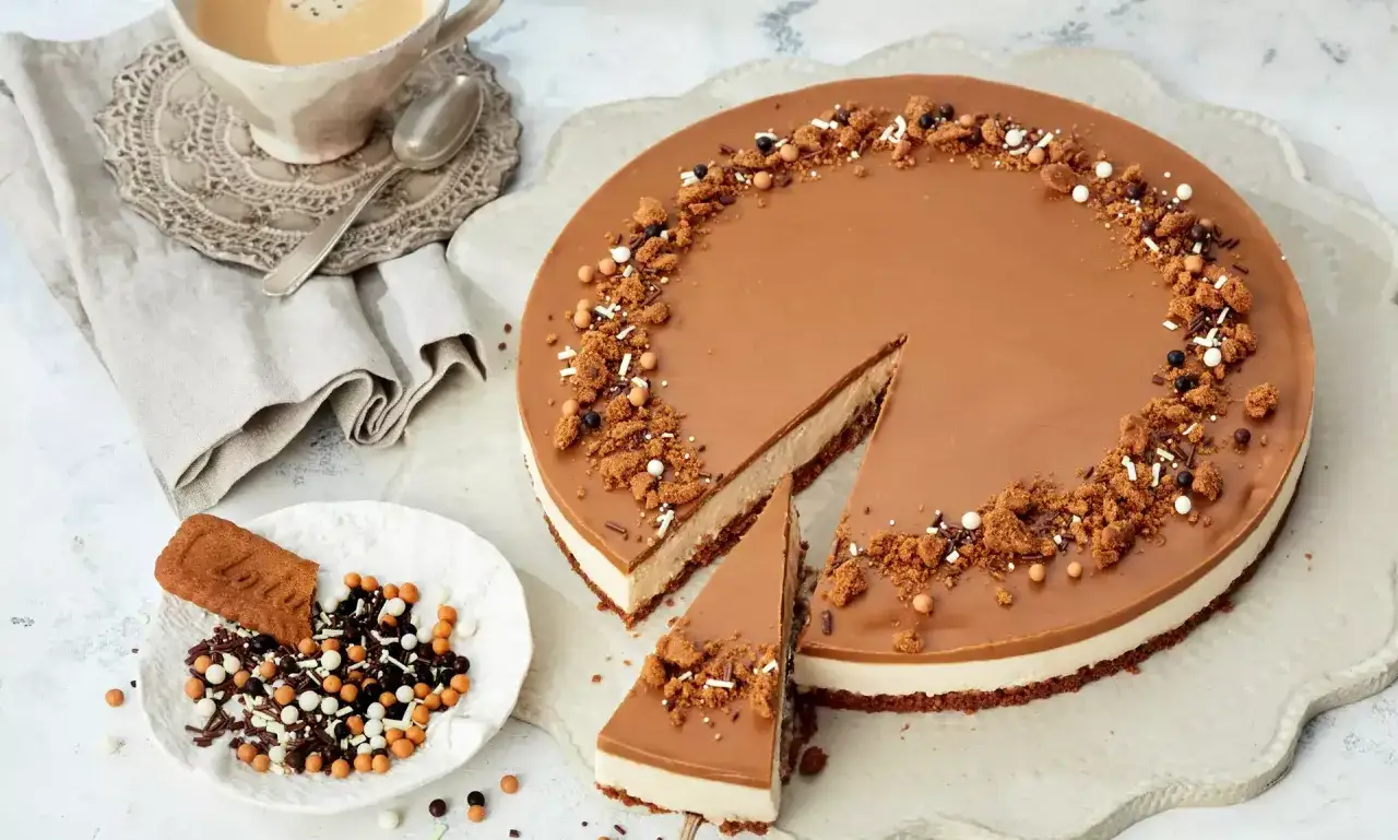 Ein köstlicher Lotus Kuchen mit Mascarpone, dekoriert mit Keksbröseln und Streuseln. Daneben eine Tasse Kaffee und ein Löffel.