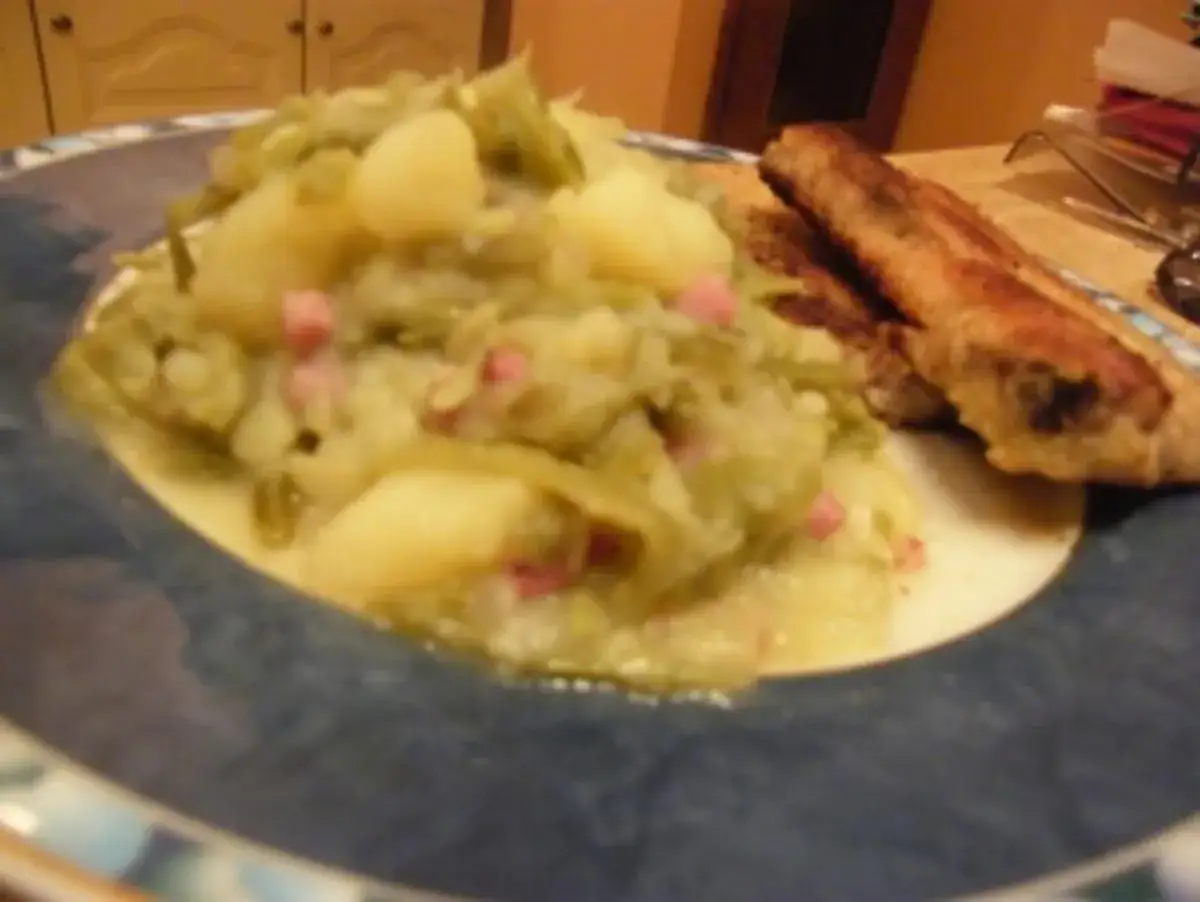 Herzhaftes Gericht mit sauren Bohnen, Kartoffeln und Speckwürfeln, dazu zwei Bratwürste auf einem blauen Teller.