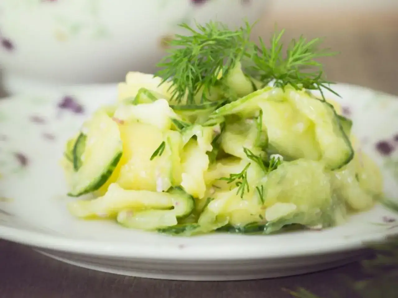 Ein cremiger Kartoffelsalat mit frischen Gurkenscheiben und Dill garniert, serviert auf einem Teller mit zartem Blumenmuster.
