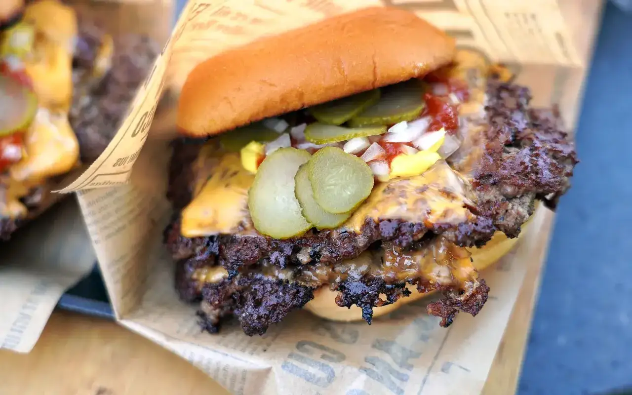 Saftiger Smash Burger mit geschmolzenem K&auml;se, Gurken, Zwiebeln und Ketchup auf einem Br&ouml;tchen, eingewickelt in Papier.