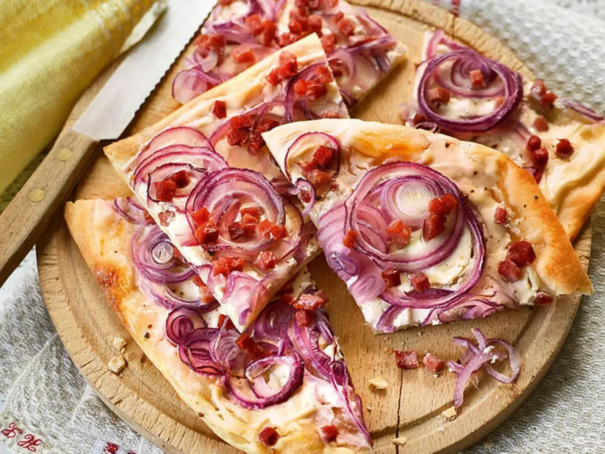 Flammkuchen Schnecken mit roten Zwiebeln und Speck auf einem Holzbrett, daneben ein Messer.