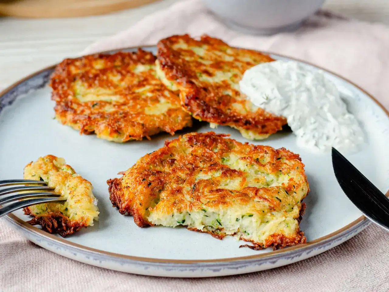 Goldbraune Zucchini-Puffer auf einem Teller, serviert mit einem Klecks Dip. Eine Gabel nimmt ein Stück eines Puffers auf.