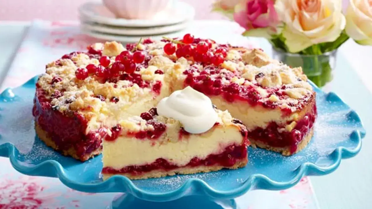 Ein saftiger Johannisbeerkuchen mit Quark und Pudding, bestreut mit Streuseln und frischen roten Johannisbeeren, serviert auf einem blauen Tortenständer.