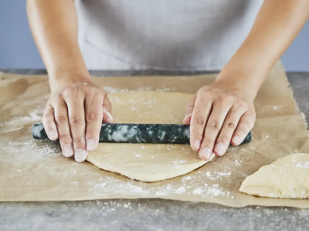 Hände rollen dünnen Flammkuchenteig auf Backpapier aus.
