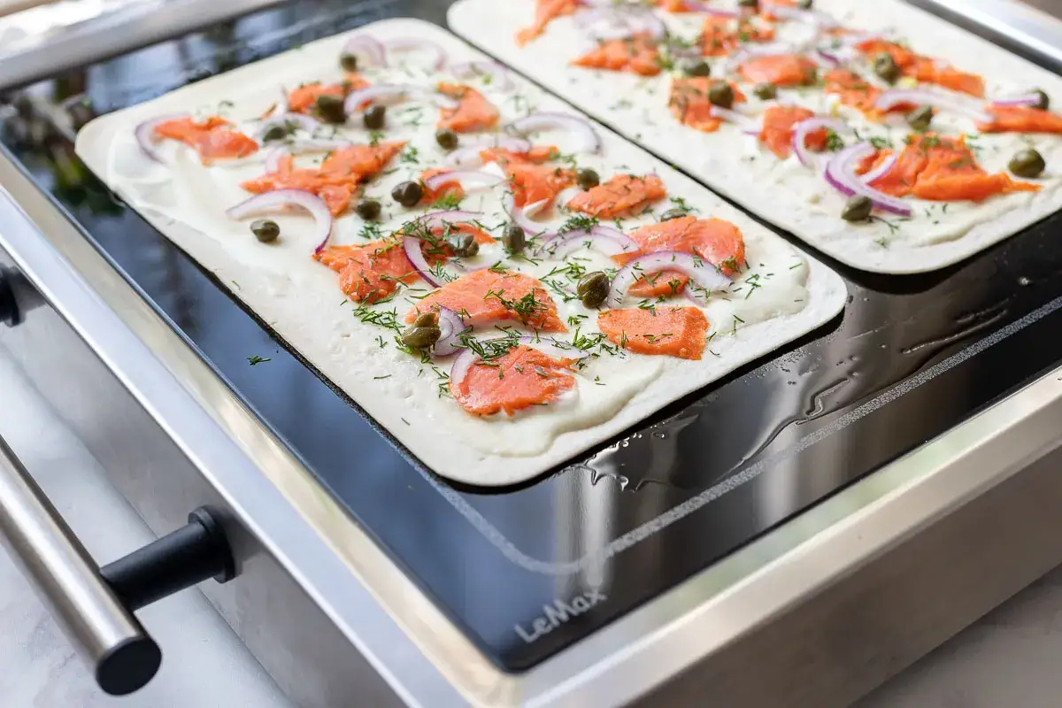 Zwei Flammkuchen mit Lachs, roten Zwiebeln und Kapern werden auf einem hei&szlig;en Grill zubereitet.