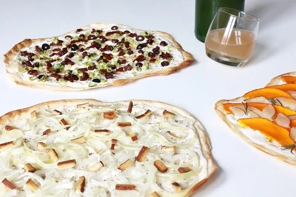 Drei Flammkuchen: einer mit Zwiebeln und Speck, ein vegetarischer Flammkuchen mit Tomaten und Oliven, und ein weiterer mit K&uuml;rbis.