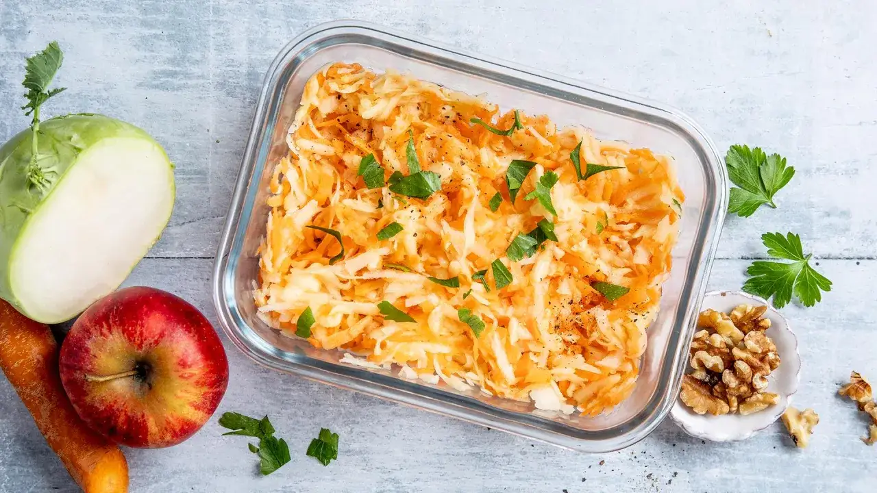 Frischer Kohlrabi-Salat mit Karotten und Apfel, garniert mit Petersilie. Daneben liegen ein halber Kohlrabi, eine Karotte, ein Apfel und Waln&uuml;sse.