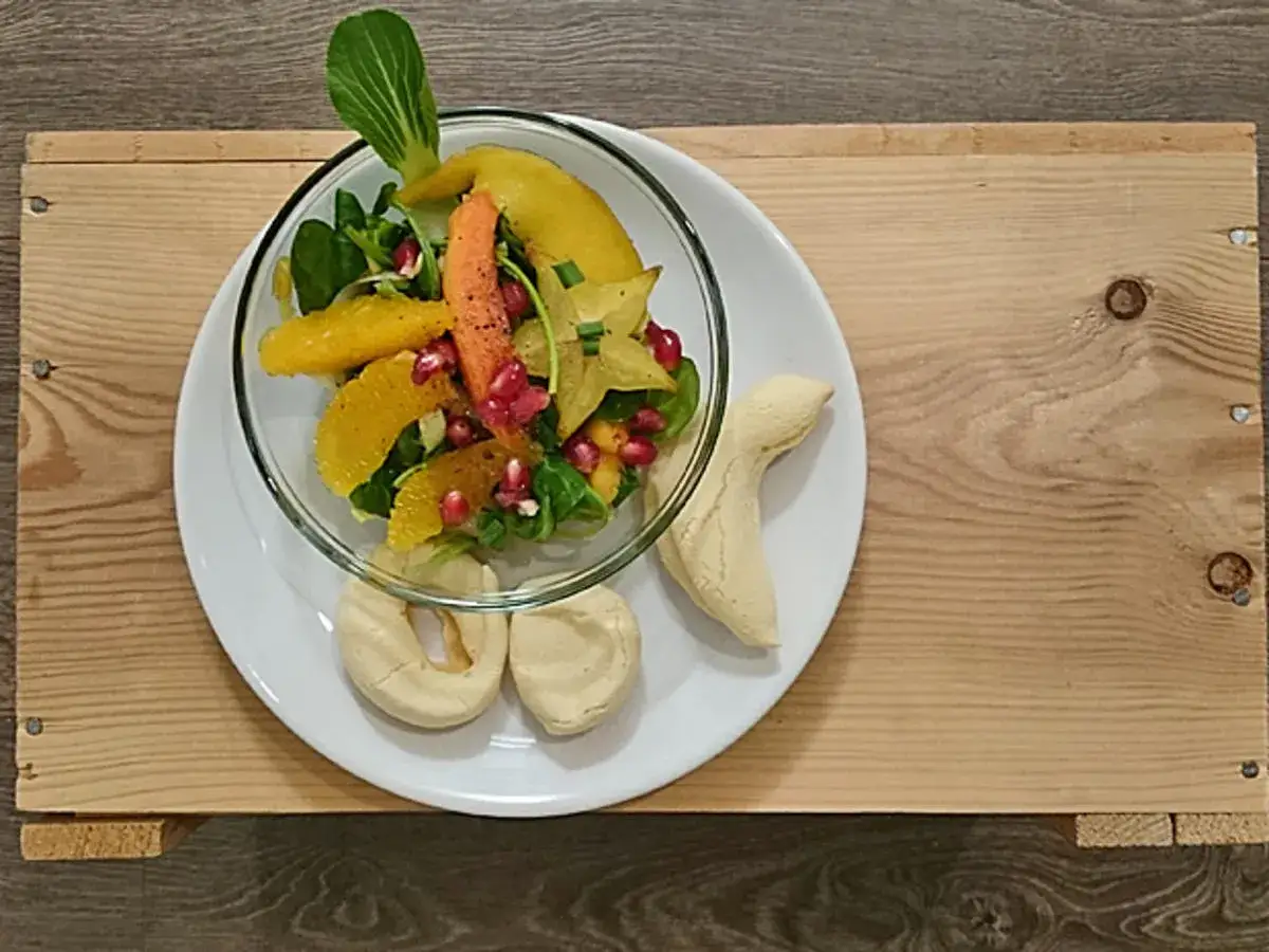 Leichte Salate als Vorspeise: Ein bunter Obstsalat mit Orangen, Granatapfel und Sternfrucht, serviert mit Geb&auml;ck auf einem Holzteller.