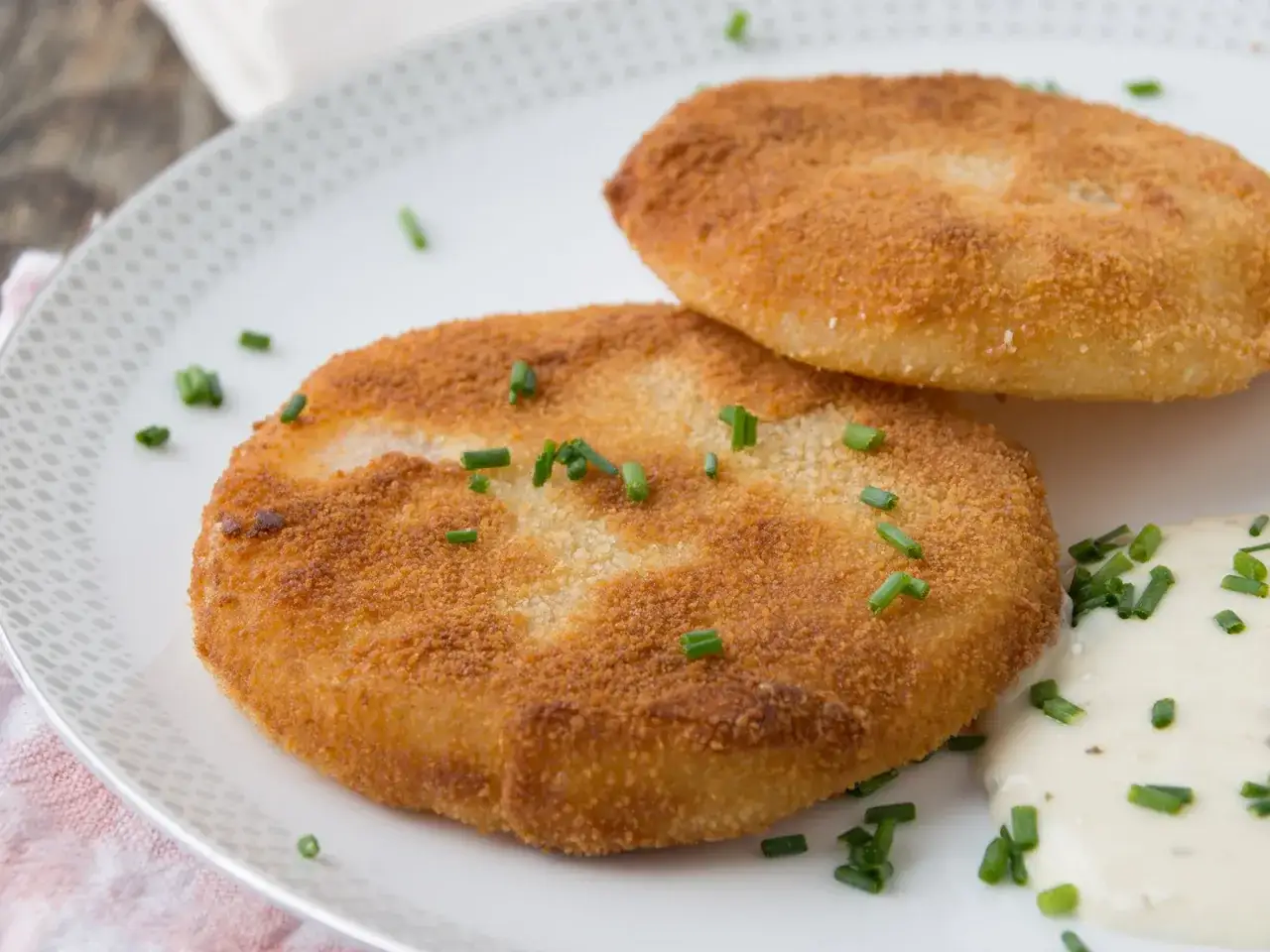 Zwei goldbraune Kohlrabi-Schnitzel, paniert und mit Schnittlauch bestreut, serviert mit einer cremigen Sauce.
