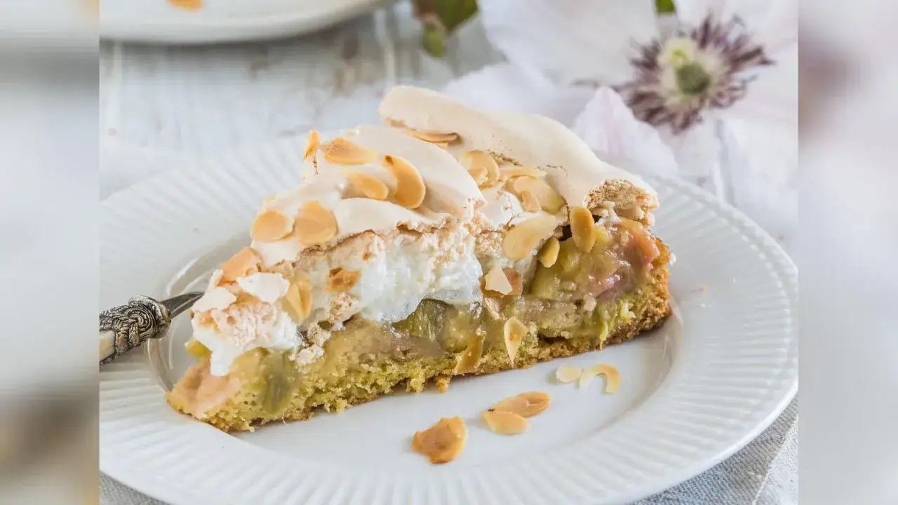Ein Stück Rhabarberkuchen mit Baiser und Mandelblättchen auf einem weißen Teller.