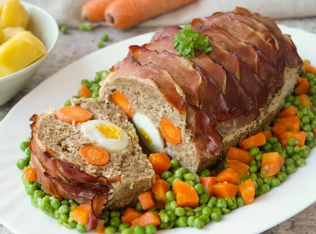 Ein herzhafter falscher Hase, umhüllt von Speck, mit Karotten und Eiern gefüllt, serviert auf Erbsen und Karotten.