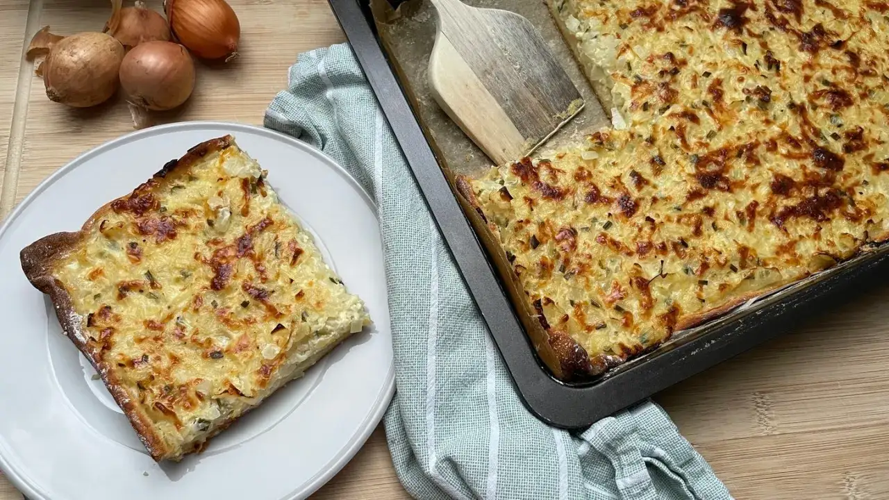 Ein frisch gebackener Zwiebelkuchen, der nach einem einfachen Rezept zubereitet wurde, liegt auf einem Blech. Eine Portion ist bereits auf einem Teller angerichtet.
