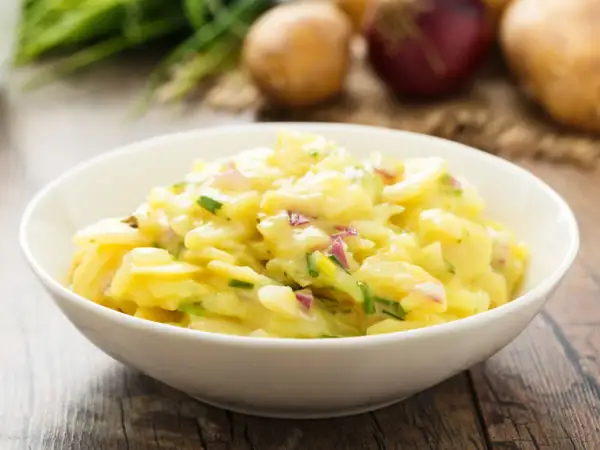 Ein Schüsselchen mit Omas Kartoffelsalat, cremig und mit frischen Kräutern verfeinert. Dazu liegen Kartoffeln und Zwiebeln bereit.