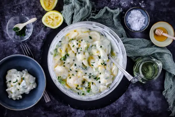 Cremiger Kartoffelsalat mit Joghurt, garniert mit Schnittlauch. Dazu Zitronenspalten und eine kleine Schüssel mit Kräutern.