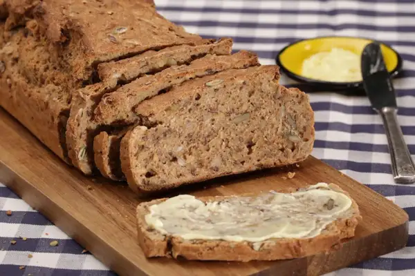 Ein frisch gebackenes Vollkornbrot mit vielen Kernen, aufgeschnitten auf einem Holzbrett. Ein Stück ist mit Butter bestrichen. Perfekt für ein gesundes Frühstück oder Abendbrot.