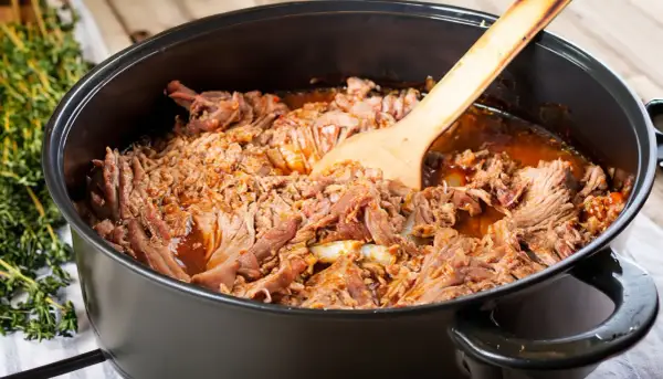 Zartes pulled pork im Dutch Oven, bereit zum Servieren. Ein Holzlöffel liegt darin.