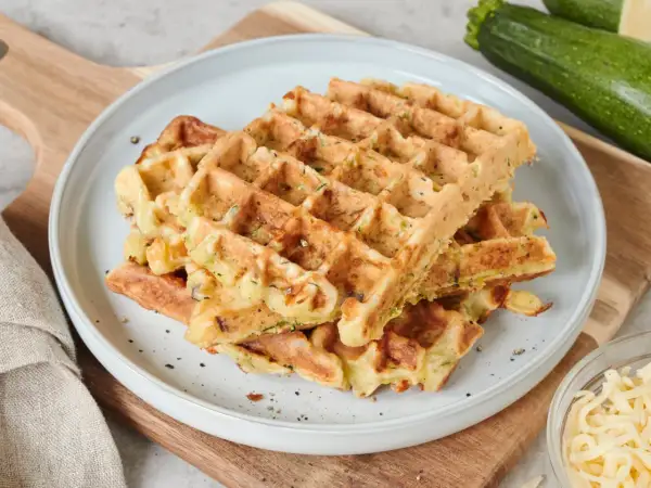 Goldbraune Zucchini-Waffeln auf einem Teller, daneben frische Zucchini und geriebener Käse. Ein leckeres zucchini waffeln rezept.