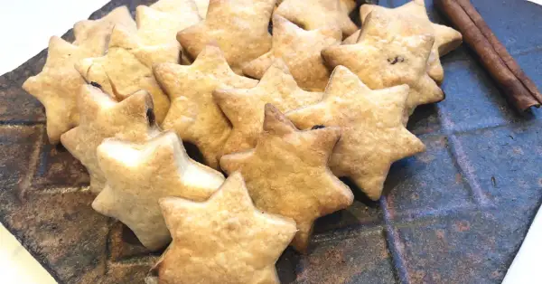 Ein Haufen goldbrauner Dinkelkekse ohne Zucker in Sternform, daneben Zimtstangen auf einem rustikalen Teller.