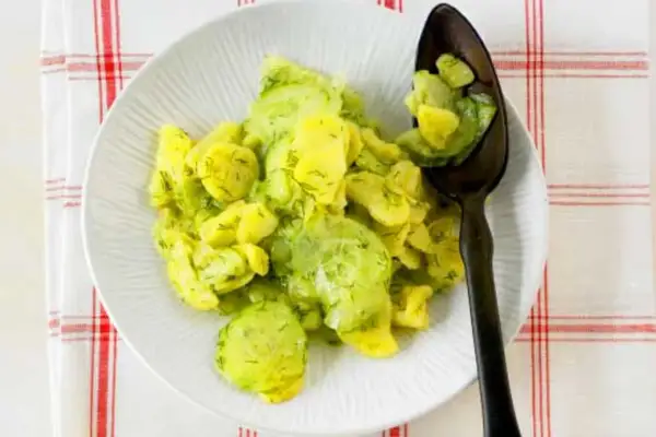 Ein Teller mit Kartoffelsalat mit Gurken und Dill, garniert mit frischem Dill. Ein Löffel mit dem Salat liegt auf dem Teller.