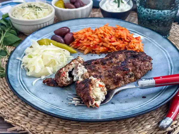 Gefülltes Bifteki-Rezept auf einem Teller mit Reisnudeln, Krautsalat, Oliven und Peperoni. Ein Stück wird mit einer Gabel angeschnitten.