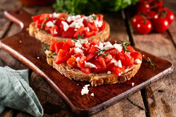Zwei rustikale Scheiben Brot belegt mit frischen Tomatenwürfeln und zerbröseltem Feta. Bruschetta mit Feta, garniert mit Kräutern, auf einem Holzbrett.