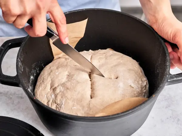 Hände schneiden mit Messer in den Teig für ein "friss dich dumm Brot" in einem Gusseisentopf.