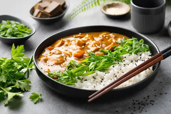 Ein Teller mit dampfendem japanischem Curry, Reis, Frühlingszwiebeln und Petersilie. Daneben kleine Schalen mit Currywürfeln und Gewürzen.