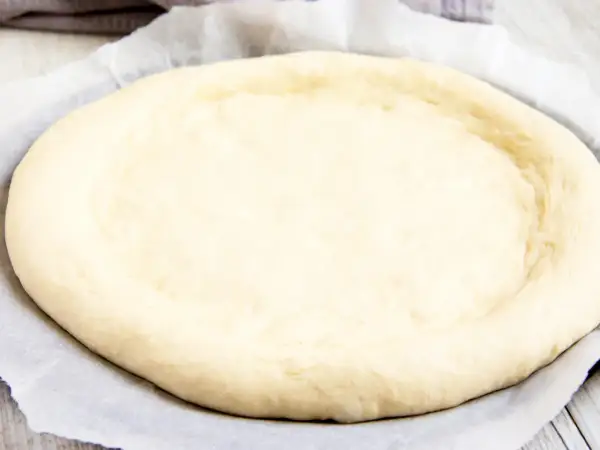 Ein runder, heller pizzateig mit trockenhefe liegt auf Backpapier. Die Ränder sind leicht angehoben.