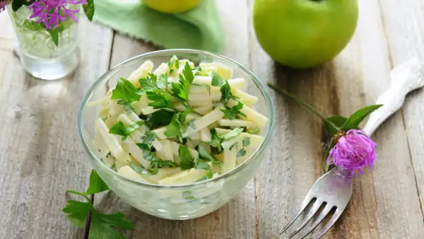 Frischer Kohlrabi Salat mit Petersilie in einer Glasschüssel, garniert mit einer lila Blüte und grünen Äpfeln im Hintergrund.